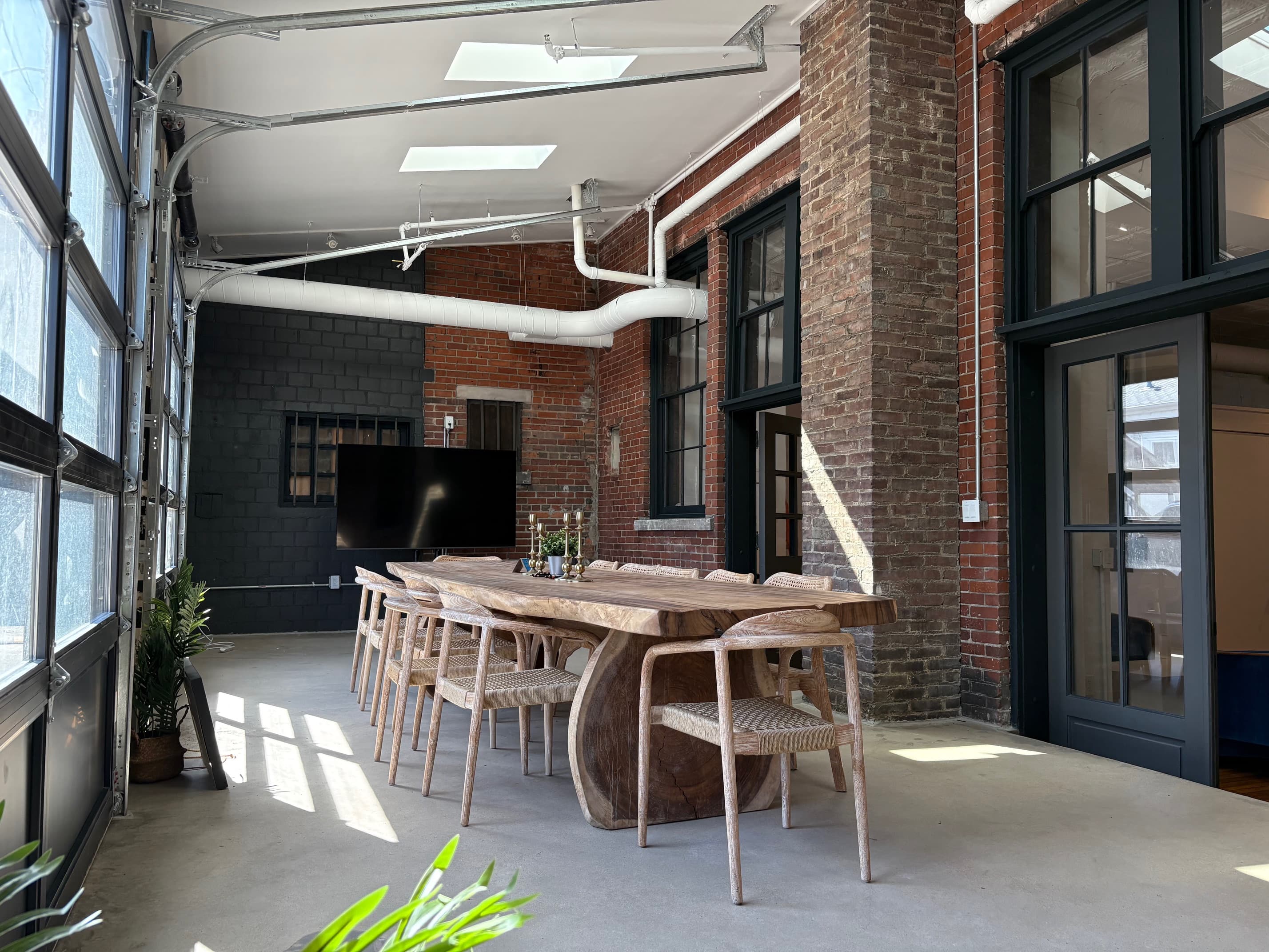 Event space with live-edge wood table, woven chairs, garage-style windows, and exposed brick